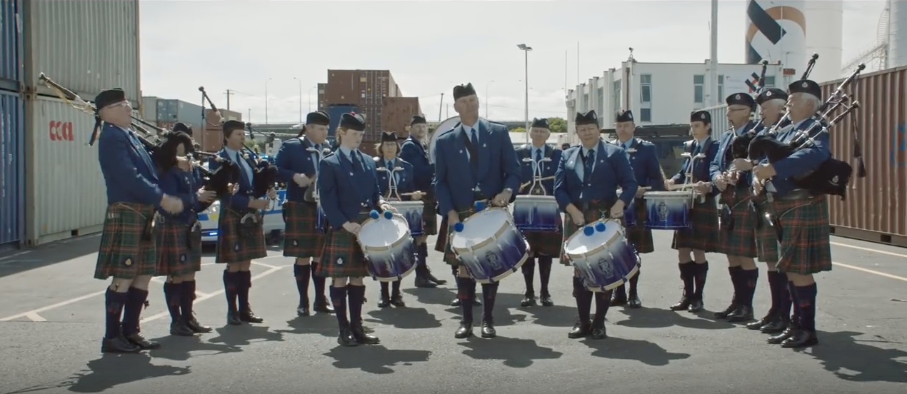 Image of men in kilts playing bagpipes and playing drums, distracting the female officer and her police team