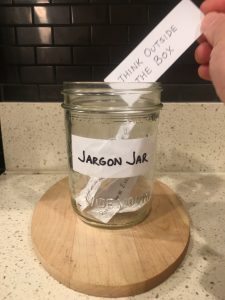 Image of hand depositing a piece of paper that reads think outside the box into the open Jargon Jar